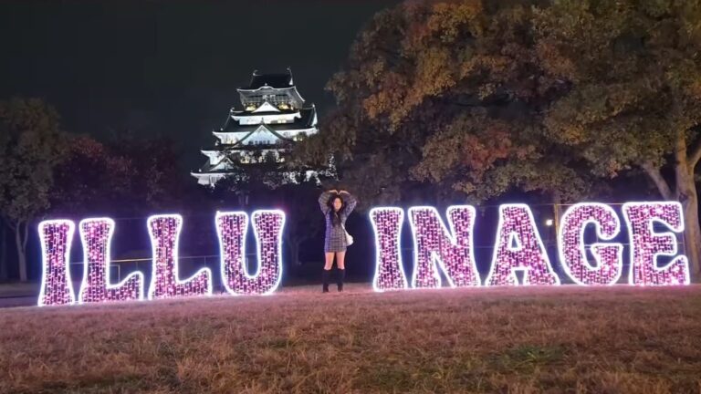 Hazu in front of the Illuminage sign doing a heart sign with her hands with Osaka castle in the background.